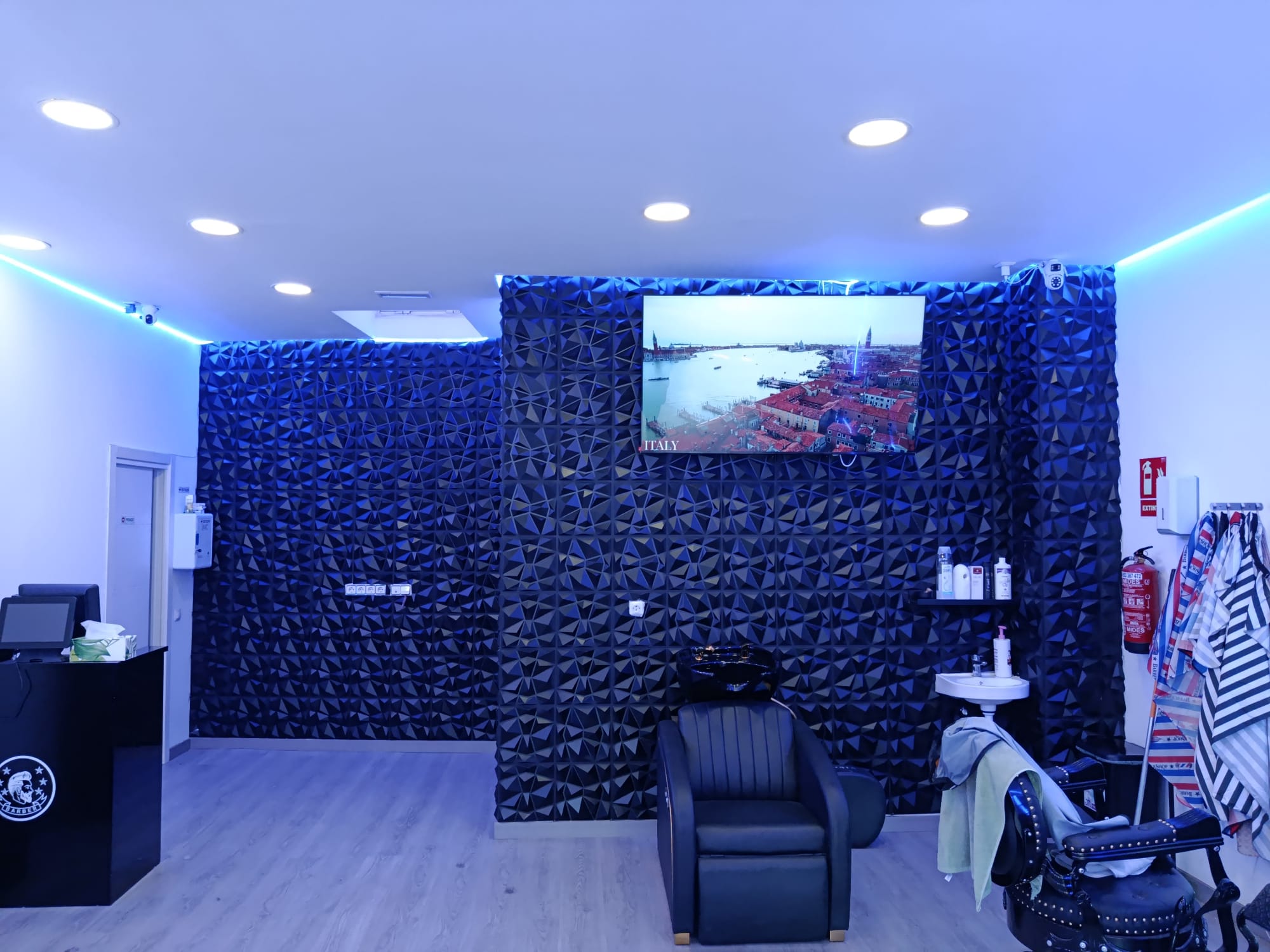 Interior de Ibra & Nor: pared geométrica negra, televisión, sillones de barbero y luz azul.